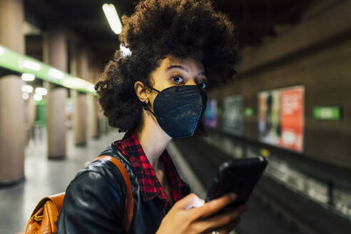 Junge Frau mit Schutzmaske hält Handy in der U-Bahn - MEUF03388
