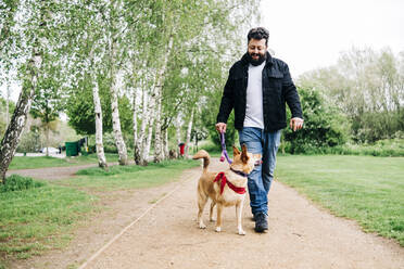 Smiling man walking with dog on footpath at park - ASGF00764
