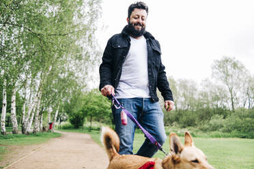 Man holding leash while walking with dog on footpath at park - ASGF00765