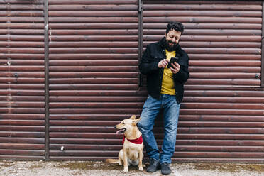 Man using smart phone by dog while leaning on brown wooden wall - ASGF00768