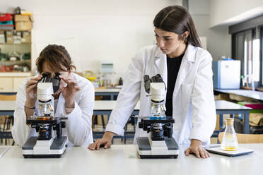 Young female scientist looking at mature colleague analyzing through microscope in laboratory - DLTSF01974