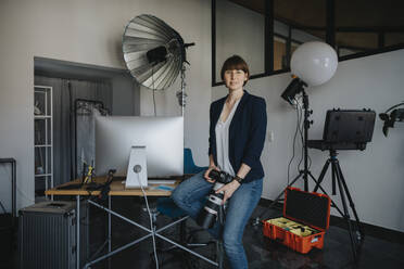 Confident female photographer holding digital camera while sitting on desk at studio - MFF08248