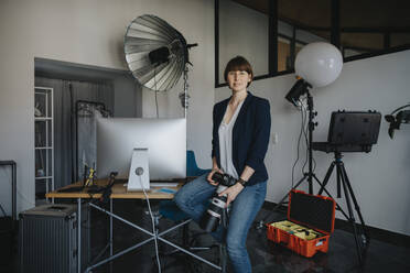 Selbstbewusste Fotografin, die eine Digitalkamera hält, während sie im Studio auf dem Schreibtisch sitzt - MFF08248