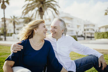 Happy mature couple looking at each other while sitting on grass during vacation - GMCF00133