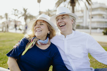 Cheerful mature couple wearing hats while sitting on grass during vacation - GMCF00139