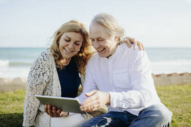 Glückliches reifes Paar macht Videoanruf durch digitale Tablet sitzen auf Gras während des sonnigen Tages - GMCF00143