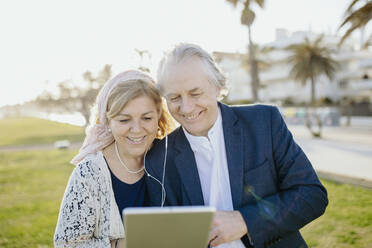 Lächelndes Paar mit In-Ear-Kopfhörern, das im Urlaub ein Video auf einem digitalen Tablet ansieht - GMCF00148