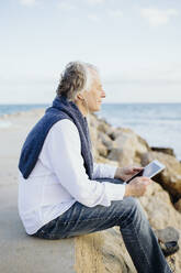 Älterer Mann hört Musik über In-Ear-Kopfhörer, während er am Meer sitzt - GMCF00154