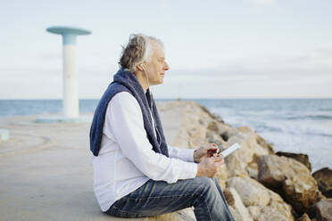 Älterer Mann sitzt mit digitalem Tablet und hört Musik am Meer - GMCF00155