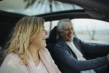 Mature blond woman laughing by man sitting in car - GMCF00163