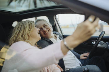 Happy mature couple looking at each other while taking selfie sitting in car - GMCF00164