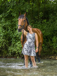 Lächelnde Frau steht mit Pferd im Fluss im Wald - STSF02987