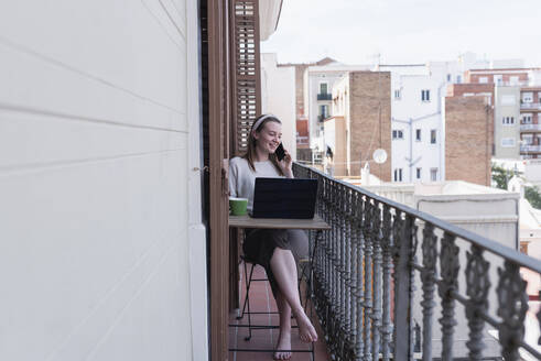 Freiberufliche Mitarbeiterin, die am Schreibtisch sitzend auf einem Balkon mit ihrem Smartphone telefoniert - DSIF00461