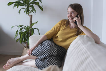 Lächelnde Frau auf dem Sofa sitzend, während sie zu Hause mit ihrem Smartphone telefoniert - DSIF00463