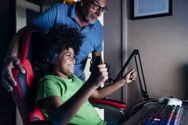 Fröhlicher Junge, der mit seinem Großvater den Sieg feiert, während er zu Hause ein Videospiel spielt - MEUF03413
