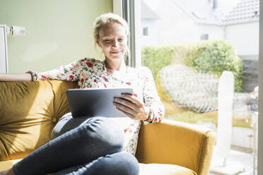 Lächelnde Frau, die ein digitales Tablet benutzt, während sie auf dem Sofa sitzt - UUF23820