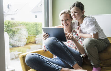 Lächelnde Mutter und Tochter benutzen ein digitales Tablet, während sie auf dem Sofa sitzen - UUF23822