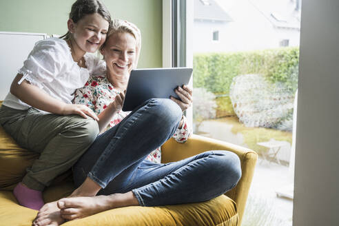 Frau benutzt digitales Tablet, während sie mit einem Mädchen zu Hause auf dem Sofa sitzt - UUF23823