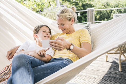 Smiling daughter looking at mother while resting in hammock at balcony - UUF23826