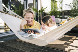Smiling mother and daughter using digital tablet while lying in hammock - UUF23830