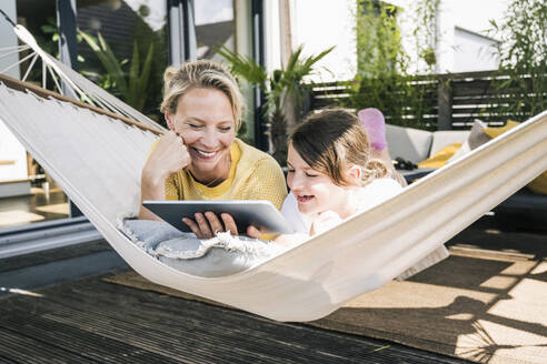 Lächelnde Mutter und Tochter benutzen ein digitales Tablet, während sie in der Hängematte liegen - UUF23830