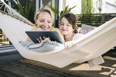 Woman and girl using digital tablet while lying in hammock at balcony - UUF23831