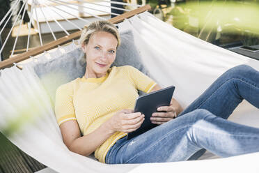 Frau mit digitalem Tablet lächelnd beim Entspannen in der Hängematte auf dem Balkon - UUF23834