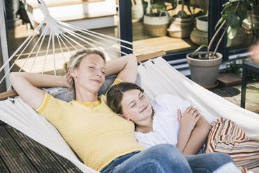 Mother with hands behind head lying by daughter in hammock at balcony - UUF23838