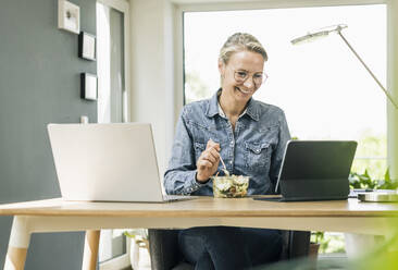 Lächelnde Geschäftsfrau beim Essen, während sie zu Hause ein digitales Tablet benutzt - UUF23841