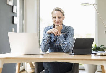Lächelnde Freiberuflerin mit Laptop und digitalem Tablet, die mit der Hand am Kinn im Home Office sitzt - UUF23845