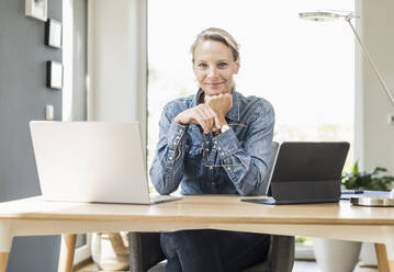 Smiling female freelancer with laptop and digital tablet sitting with hand on chin at home office - UUF23845