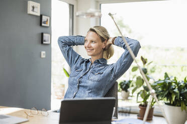 Smiling businesswoman with hands behind head sitting at home office - UUF23846