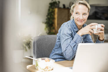 Weibliche Freiberuflerin lächelt, während sie eine Kaffeetasse im Heimbüro hält - UUF23848