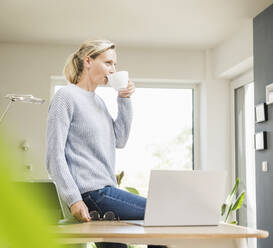 Female freelancer drinking coffee while sitting by laptop on desk - UUF23855
