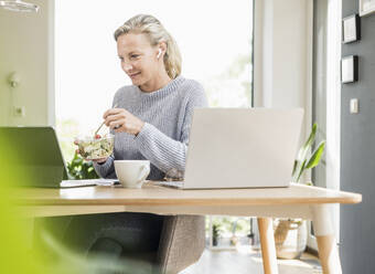 Smiling businesswoman eating food while working at home - UUF23856