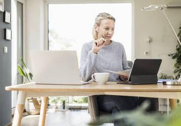 Businesswoman with digital tablet and laptop holding paper while working at home office - UUF23858