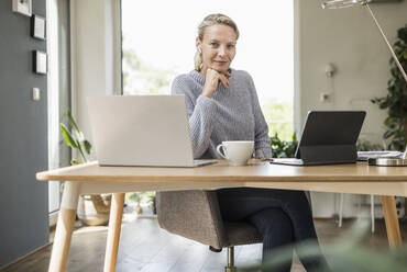 Businesswoman with hand on chin sitting by desk at home office - UUF23860