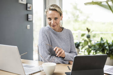Businesswoman holding eyeglasses while sitting by laptop and digital tablet at home - UUF23861