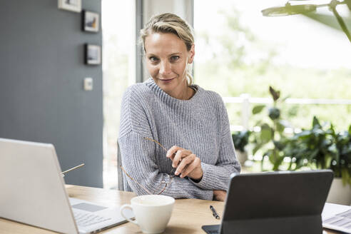 Geschäftsfrau mit Brille, die zu Hause an einem Laptop und einem digitalen Tablet sitzt - UUF23861