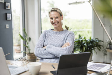 Female freelancer with arms crossed sitting at home office - UUF23862