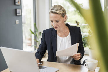 Geschäftsfrau hält ein Dokument, während sie einen Laptop im Büro benutzt - UUF23875