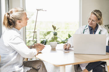 Arzt schreibt auf Papier, während er mit einem Patienten im Büro sitzt - UUF23882