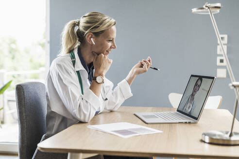 Smiling female doctor having discussion with colleague through video call on laptop at office - UUF23888