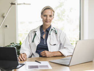 Ärztin mit Laptop und digitalem Tablet im Büro sitzend - UUF23893