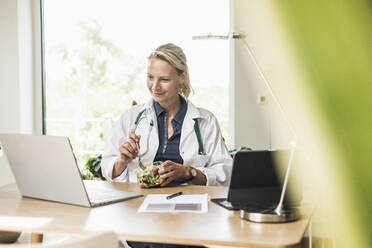 Smiling female doctor eating food while looking at laptop in office - UUF23895