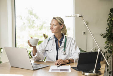 Ärztin mit Laptop, die eine Kaffeetasse hält, während sie im Büro sitzt - UUF23896