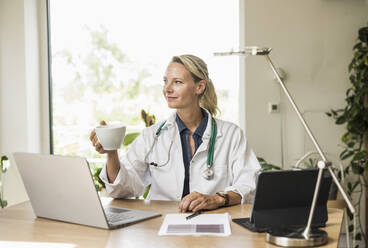 Female doctor with laptop holding coffee cup while sitting at office - UUF23896