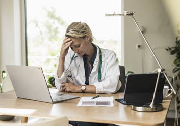 Doctor with head in hands sitting by desk at office - UUF23897