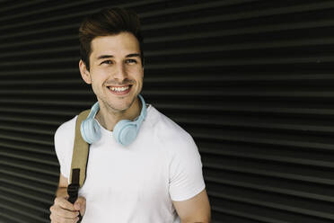 Smiling young man looking away while carrying backpack in front of shutter - XLGF02068