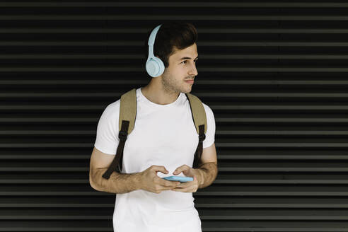 Junger Mann mit Handy schaut weg, während er vor einem Fensterladen steht - XLGF02069